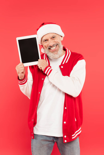 cheerful middle aged man in santa hat holding digital tablet isolated on red - Photo, Image