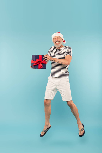 comprimento total do homem feliz de meia idade em santa chapéu saltando com caixa de presente em azul - Foto, Imagem