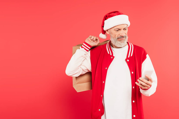 happy middle aged man in santa hat holding shopping bags and using smartphone on red - Photo, Image