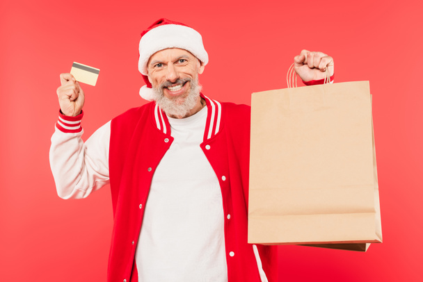 Noel Baba şapkalı mutlu orta yaşlı bir adam elinde alışveriş çantaları ve kredi kartıyla kırmızıda izole edilmiş. - Fotoğraf, Görsel