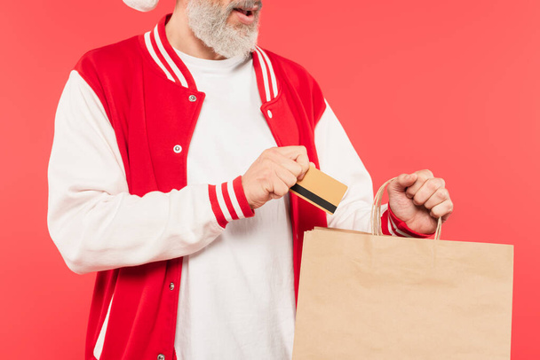 cropped view of middle aged man putting credit card in shopping bag isolated on red - Photo, Image
