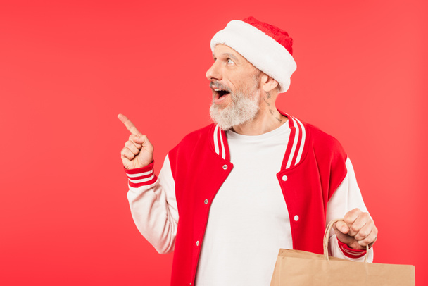 espantado homem de meia idade apontando com o dedo e segurando saco de compras isolado no vermelho - Foto, Imagem