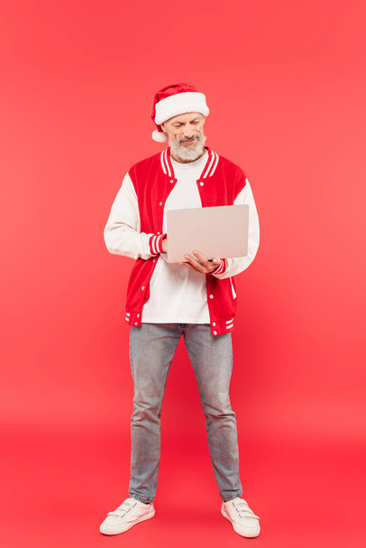 plná délka usmívající se na volné noze středního věku v Santa klobouku pomocí notebooku na červené - Fotografie, Obrázek