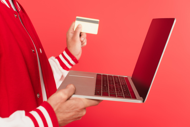 καλλιεργημένη άποψη μεσήλικας άνδρας που κρατά φορητό υπολογιστή και πιστωτική κάρτα που απομονώνονται σε κόκκινο - Φωτογραφία, εικόνα