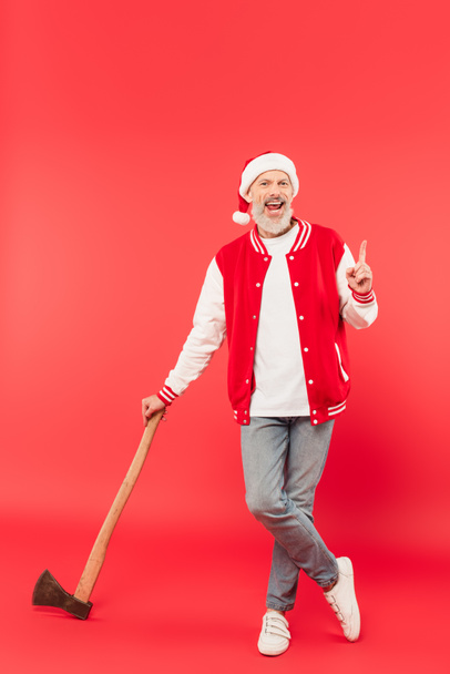 täyspitkä onnellinen keski-ikäinen mies joulupukki hattu tilalla kirves ja sormella punainen - Valokuva, kuva