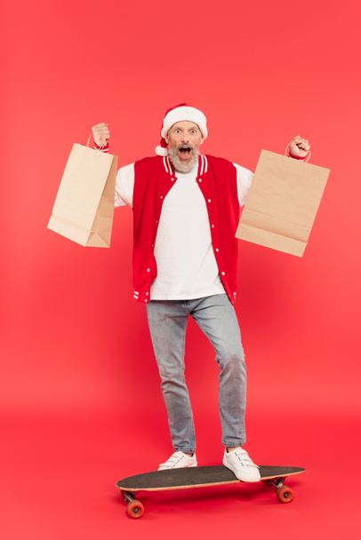 pełna długość wstrząśnięty mężczyzna w średnim wieku w kapeluszu Santa trzymając torby na zakupy podczas jazdy na deskorolce na czerwono - Zdjęcie, obraz
