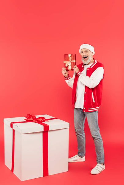 pełna długość podekscytowany mężczyzna w średnim wieku w kapeluszu Santa trzymając pudełko prezentów blisko ogromny prezent na czerwono - Zdjęcie, obraz