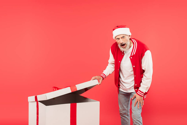 Şok olmuş orta yaşlı Noel Baba şapkalı adam kırmızı üzerine izole edilmiş büyük bir hediye açıyor. - Fotoğraf, Görsel