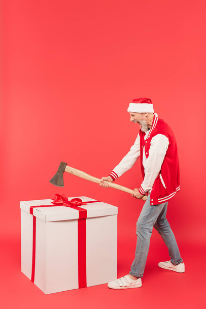 volle Länge des Mannes mittleren Alters mit Weihnachtsmannmütze, der eine Axt hält, während er in der Nähe eines riesigen Geschenks auf Rot schreit - Foto, Bild