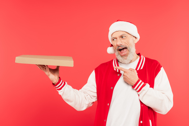 pozytywny mężczyzna w średnim wieku w Santa kapelusz wskazując na pudełko pizzy izolowane na czerwono - Zdjęcie, obraz