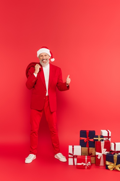 полная длина счастливый мужчина средних лет в шляпе и костюме держа Санта мешок рядом с подарками, показывая палец вверх на красный - Фото, изображение