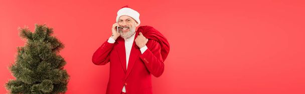happy middle aged man in hat and suit holding santa sack and having phone call near pine tree isolated on red, banner - Photo, Image