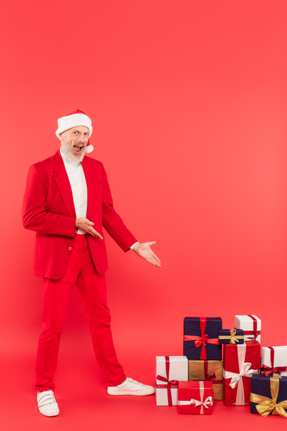 полная длина изумленный мужчина средних лет в шляпе Санты и костюм указывая руками на подарки на красный - Фото, изображение