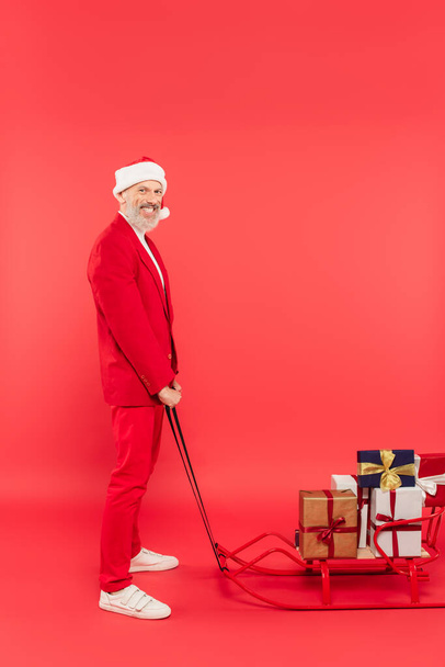 full length of cheerful middle aged man in santa hat and suit holding sleigh with presents on red - Photo, Image