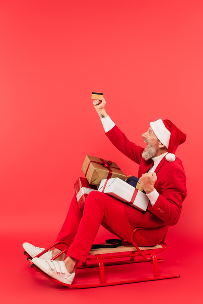 full length of happy middle aged man in santa hat and suit sitting in sleigh with presents while holding credit card on red - Photo, Image