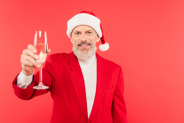 щасливий чоловік середнього віку в костюмі і капелюсі Санти тримає келих шампанського ізольовано на червоному
 - Фото, зображення
