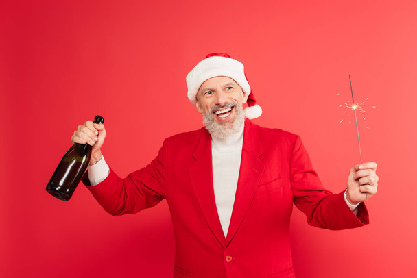 Mutlu orta yaşlı takım elbiseli adam ve elinde şampanya şişesi ve kırmızı üzerine izole edilmiş maytap tutan Noel Baba şapkalı adam. - Fotoğraf, Görsel