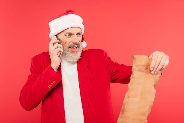 positive mature man in santa hat reading letter on craft paper while talking on smartphone isolated on red - Photo, Image