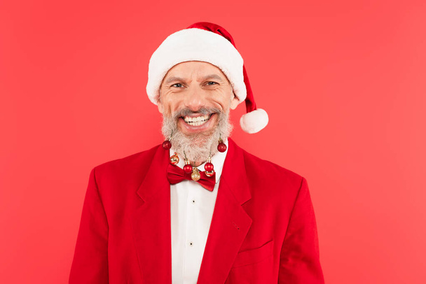 happy mature man in santa hat with christmas balls on beard isolated on red - Photo, Image