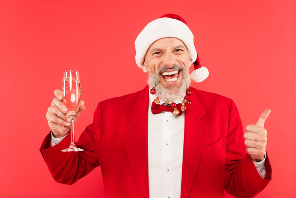hombre maduro feliz en sombrero de santa con bolas de Navidad en la barba con champán y mostrando el pulgar hacia arriba aislado en rojo - Foto, imagen