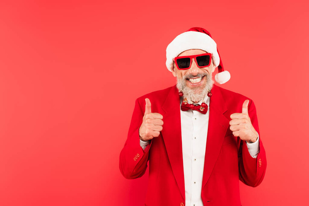 Noel Baba şapkalı ve güneş gözlüklü mutlu olgun bir adam sakallı Noel topları kırmızı renkte izole edilmiş baş parmağını gösteriyor. - Fotoğraf, Görsel