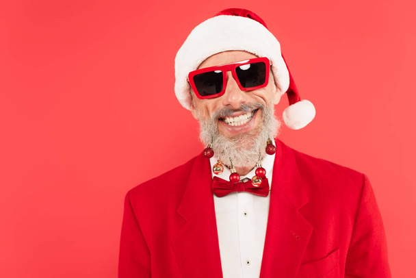 szczęśliwy Dojrzały mężczyzna w kapeluszu Mikołaja i okulary przeciwsłoneczne z kulkami świątecznymi na brodzie izolowane na czerwono - Zdjęcie, obraz