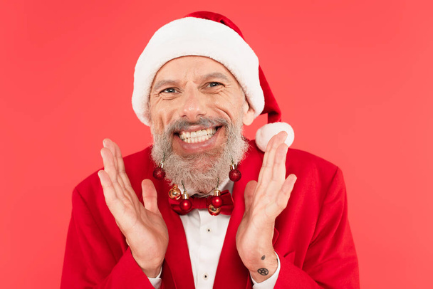excited mature man in santa hat with christmas balls on beard gesturing isolated on red - Photo, Image