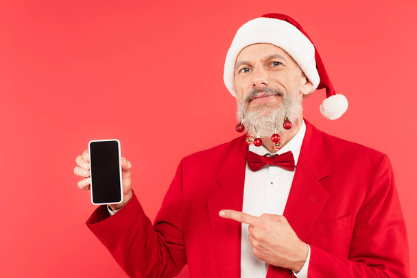 glücklich erwachsener Mann mit Weihnachtsmütze und Weihnachtskugeln am Bart zeigt mit dem Finger auf Smartphone isoliert auf rot - Foto, Bild