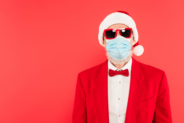 mature man in santa hat, medical mask and sunglasses isolated on red - Photo, Image