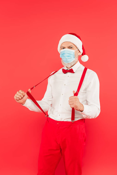 ώριμος άνδρας με καπέλο santa και ιατρική μάσκα ρύθμισης τιράντες που απομονώνονται σε κόκκινο - Φωτογραφία, εικόνα