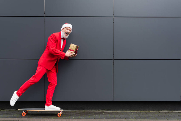サンタ・ハットの幸せな成熟した男がプレゼントを保持し、グレーの壁の近くのスケートボードに乗って - 写真・画像