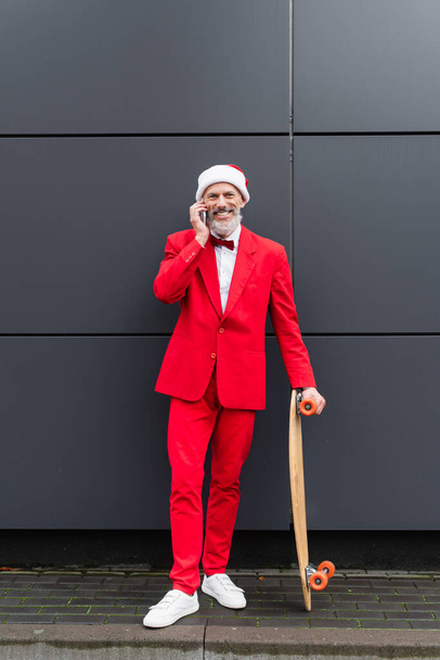 pełna długość szczęśliwy Dojrzały mężczyzna w Santa kapelusz mówi przez telefon komórkowy stojąc z deskorolką w pobliżu szarej ściany - Zdjęcie, obraz