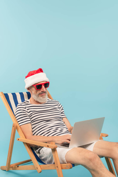 hombre maduro feliz en sombrero de santa y gafas de sol sentado en la silla de cubierta y el uso de ordenador portátil aislado en azul - Foto, Imagen