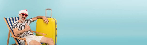 hombre maduro feliz en sombrero de santa y gafas de sol sentado en la silla de cubierta cerca de equipaje y mostrando el pulgar hacia arriba en azul, bandera - Foto, Imagen