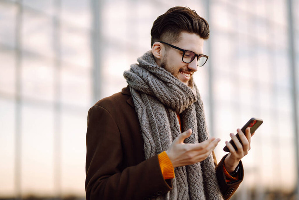 nline communication. Young man wearing  coat with a smartphone in his hand, walking in the street. Young bearded guy with modern hairstyle in urban background  typing on cell phone.  - Foto, afbeelding