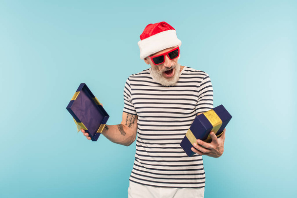 здивований чоловік середнього віку в капелюсі Санта та сонцезахисних окулярах, що відкривають подарункову коробку ізольовано на синьому
 - Фото, зображення