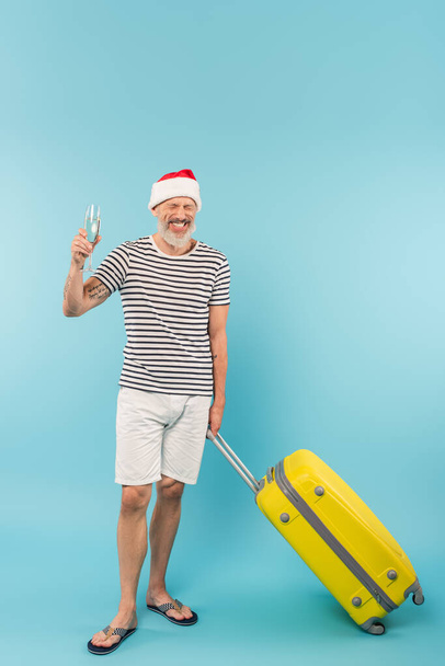 pozytywny mężczyzna w średnim wieku w Santa kapelusz stojący z bagażem i trzymając szklankę szampana na niebiesko - Zdjęcie, obraz