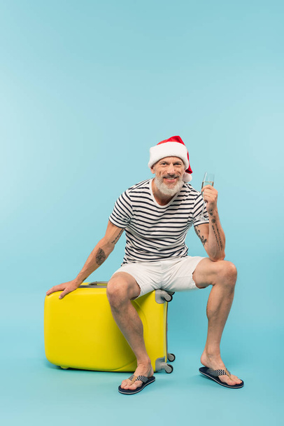fröhlicher Mann mittleren Alters mit Weihnachtsmütze sitzt auf dem Gepäck und hält ein Glas Champagner auf blauem Grund - Foto, Bild