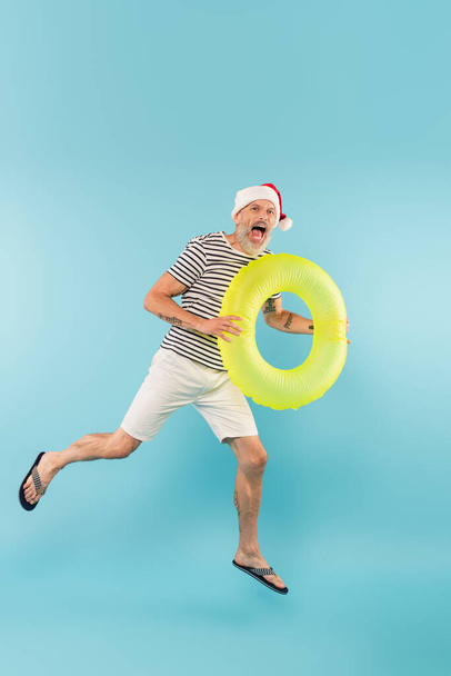 full length of middle aged man in santa hat holding inflatable ring and screaming on blue - Photo, Image