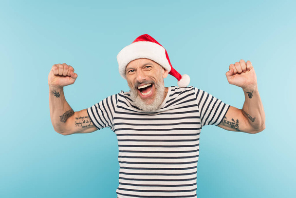 podekscytowany mężczyzna w średnim wieku w stiped t-shirt i Santa kapelusz odizolowany na niebiesko  - Zdjęcie, obraz