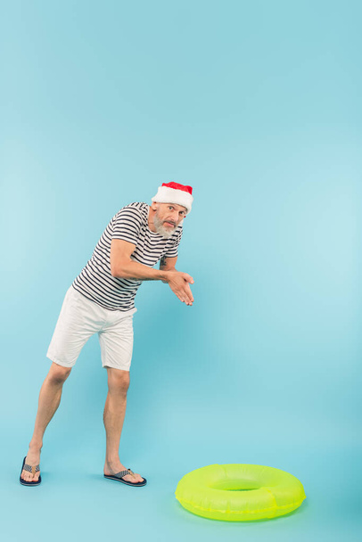 full length of middle aged man in stiped t-shirt and santa hat imitating diving near swim ring on blue - Photo, Image