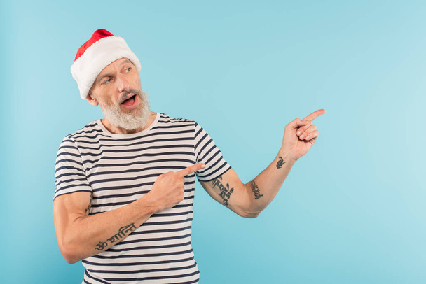 bearded and middle aged man in stiped t-shirt and santa hat pointing with fingers isolated on blue - Photo, Image