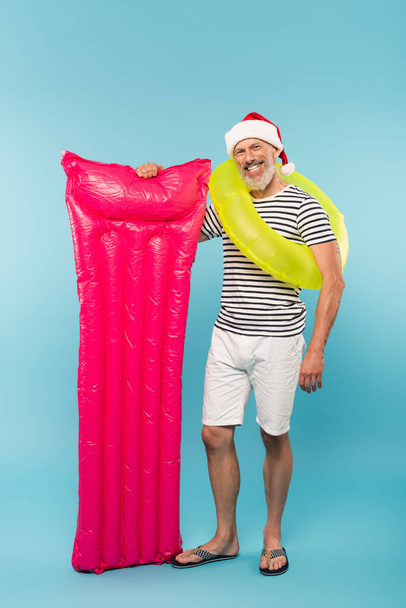 full length of happy middle aged man in santa hat and swim ring holding pink inflatable mattress on blue - Photo, Image
