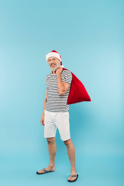 volle Länge des glücklichen Mannes mittleren Alters in Weihnachtsmütze mit Sack auf blauem Grund - Foto, Bild