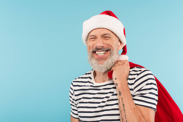 glücklicher Mann mittleren Alters mit Weihnachtsmannmütze und Sack auf blauem Grund - Foto, Bild