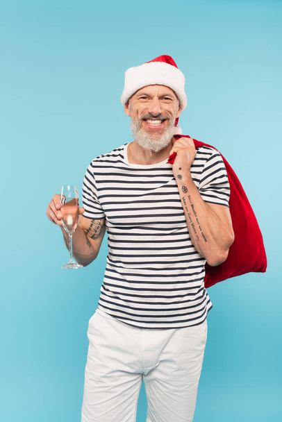 heureux homme d'âge moyen dans santa chapeau tenant sac et verre de champagne isolé sur bleu - Photo, image