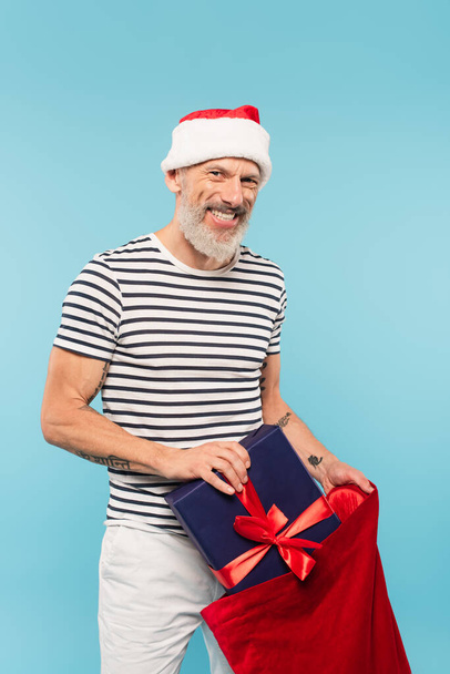 happy middle aged man in santa hat holding present and sack isolated on blue - Photo, Image