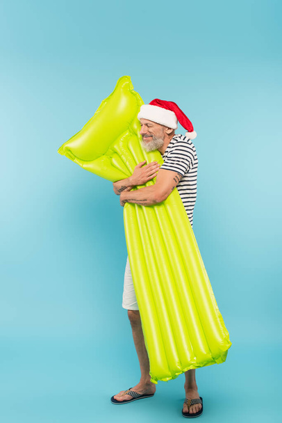 volle Länge des glücklichen Mannes mittleren Alters in Weihnachtsmannmütze umarmt aufblasbare Matratze auf blau - Foto, Bild
