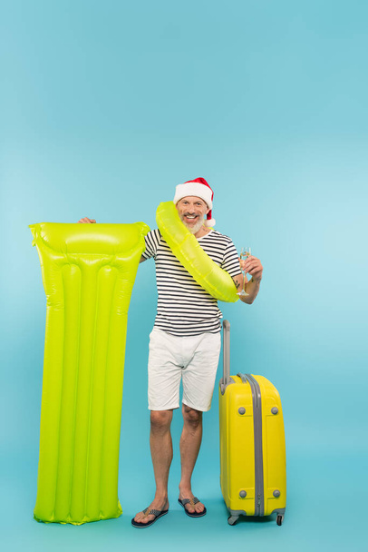 happy man in santa hat with swim ring standing near inflatable mattress, luggage and holding glass of champagne on blue - Photo, Image