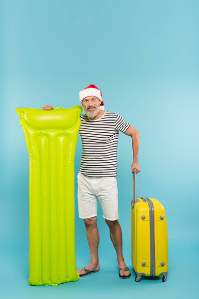 ώριμος άνδρας σε καπέλο santa στέκεται κοντά φουσκωτό στρώμα με αποσκευές σε μπλε - Φωτογραφία, εικόνα